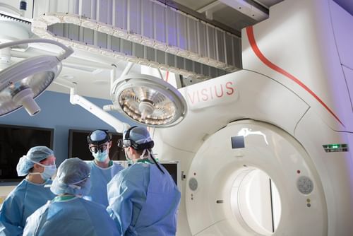 A team of medical professionals in scrubs and surgical caps focused on a VISIUS Surgical Theatre system, standing around a high-tech imaging device in a modern operating room