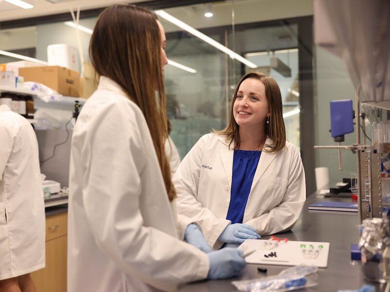 Professor Katie Hixon smiles at a graduate student. Both are wearing white lab coats and working on research in her lab.