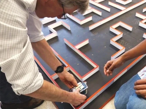 A person adjusts a small robotic mouse inside a large, complex maze with orange-bordered walls on a dark floor.