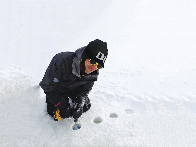 A person in winter gear and a black beanie kneels in the snow, using a tool to drill small holes in the ice.